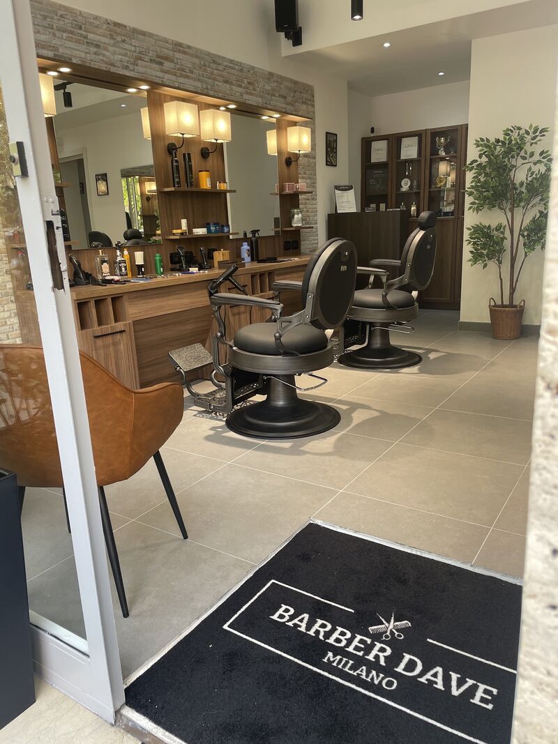 L'ingresso di Barber Dave Milano: il tappeto nero con il logo, le poltrone da barbiere nere e la parete attrezzata in legno con le lampade accese