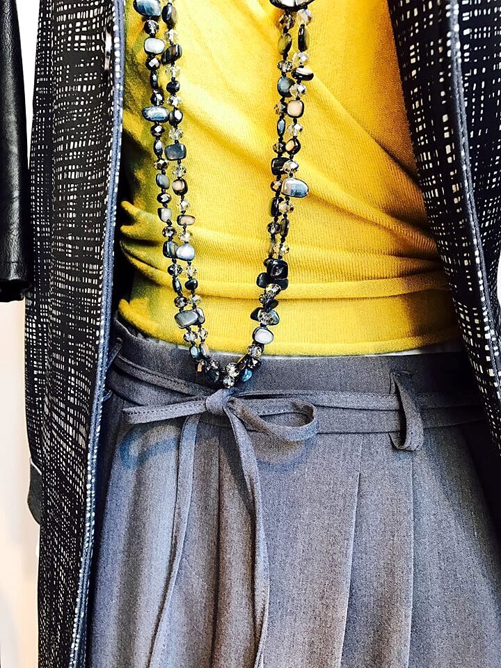 Dettaglio di collane lunghe su top giallo e pantalone grigio con fiocco in vita